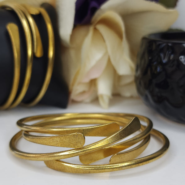 A set of gold-colored brass bangles displayed in a stacked arrangement with a blurred background featuring floral elements and other jewelry.